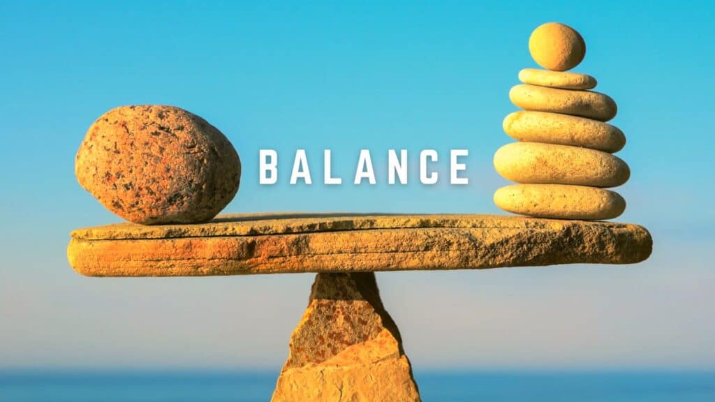 A large, rough stone on the left is perfectly balanced against a stack of seven smooth, round pebbles on the right, all resting on a flat slab supported by a single pointed rock. The word “BALANCE” appears in bold white letters against a clear blue sky and ocean horizon.