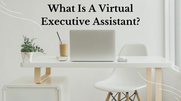 A minimalist white desk setup with a closed laptop, iced coffee, wireless mouse, and a small potted plant, set against a bright window. The text “What Is A Virtual Executive Assistant?” appears above the workspace in bold black font.