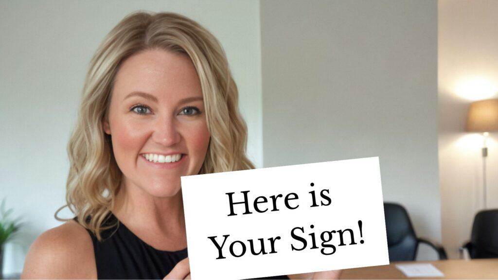 A smiling woman with blonde hair holds up a white sign that reads “Here is Your Sign!” in bold black text. She stands in a well-lit office space, creating a lighthearted and motivational vibe.