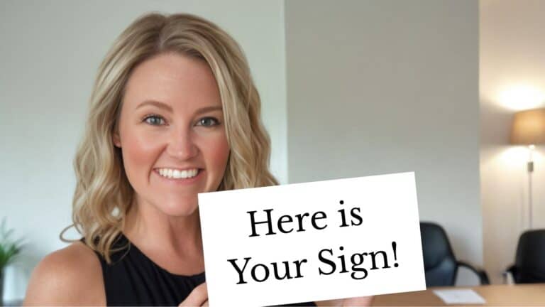A smiling woman with blonde hair holds up a white sign that reads “Here is Your Sign!” in bold black text. She stands in a well-lit office space, creating a lighthearted and motivational vibe.