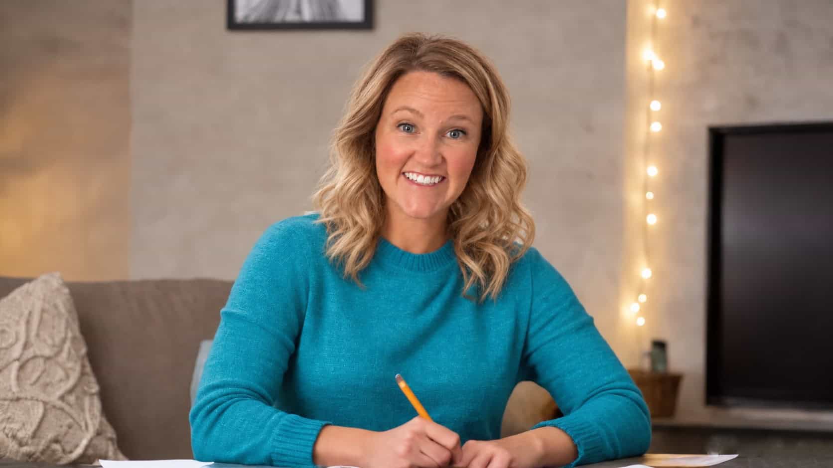 Morgan Middleton, a Certified AI Integrator and experienced Executive Assistant, smiling while writing with a pencil at a table. She’s wearing a teal sweater and seated in a cozy living room with soft lighting and string lights in the background.