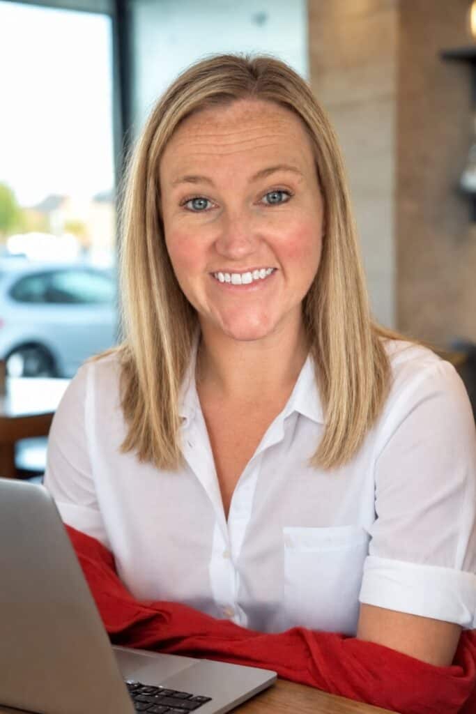 Smiling woman with shoulder-length blonde hair and blue eyes, wearing a white button-down shirt and sitting at a table with a laptop, identified as Certified AI Integrator and experienced Executive Assistant Morgan Middleton. Natural light from a window highlights her face in a cozy café setting.